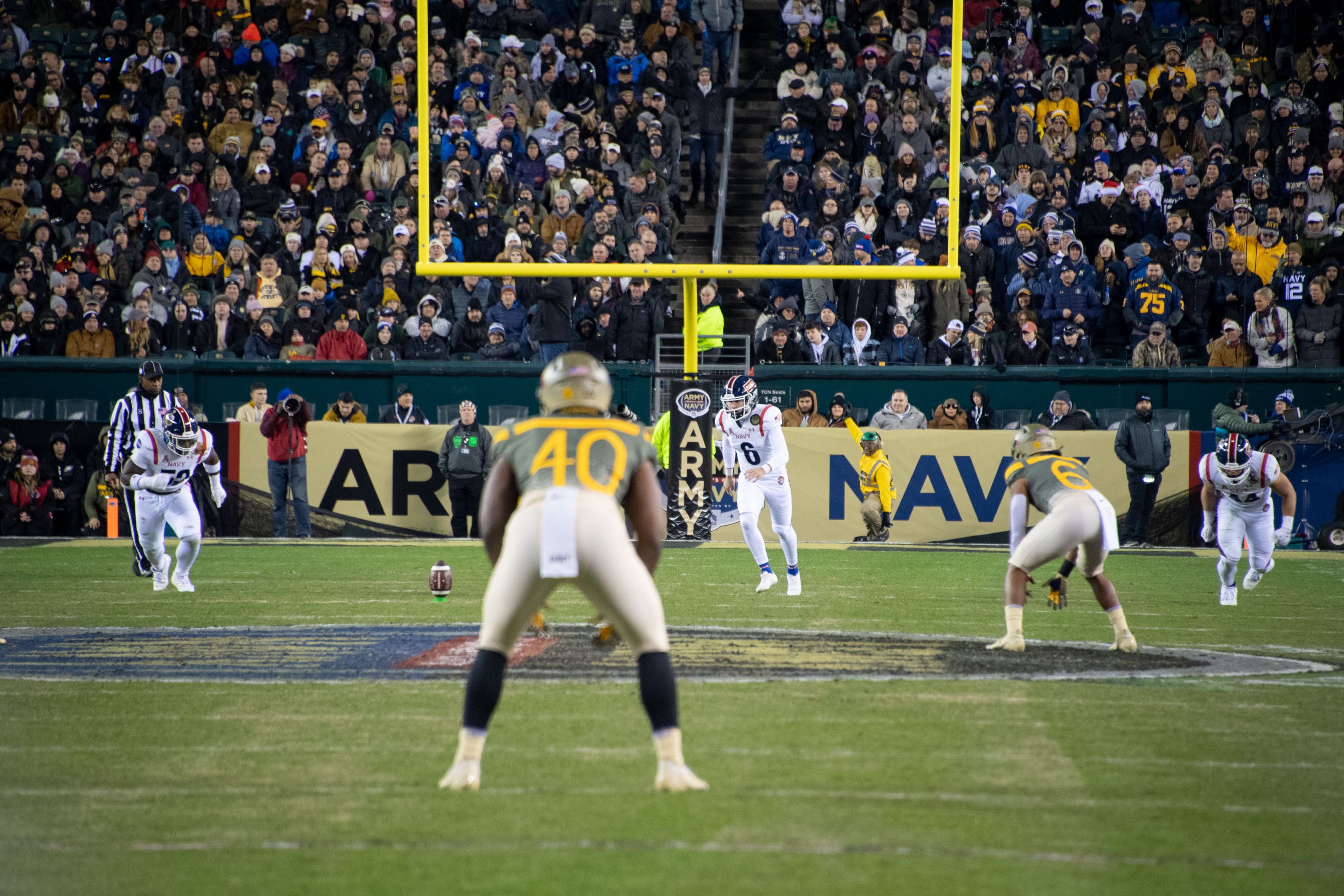 Ein Fußballspiel im Gange mit Spielern in Helmen und Sportbekleidung im Vordergrund und einer Zuschauermenge im Hintergrund, mit sichtbaren Pfählen und Tafeln mit Text.