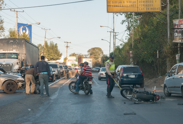 Eine Gruppe von Menschen steht um ein verunglücktes Motorrad auf der Seite einer Straße mit mehreren Fahrzeugen, darunter ein Lastwagen, und einer Hintergrund von Bäumen, Polen, Lampen und Schildern unter dem Himmel.