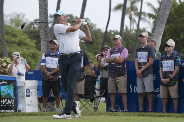 Ein Mann in einem weißen T-Shirt, schwarzen Hosen und einer blauen Mütze schwingt einen Golfschläger auf einem grünen Feld, mit Zuschauern und Bäumen im Hintergrund unter einem klaren blauen Himmel.