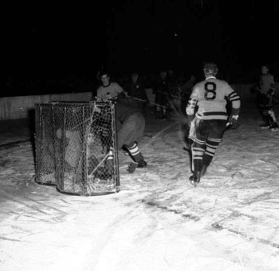 Schwarzes und weißes Foto von Männern, die Hockey auf einem Eisplatz mit einem Netz im Vordergrund und einer Wand im Hintergrund spielen.