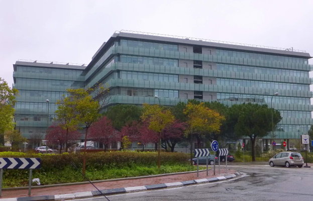 Großes modernes Gebäude mit zahlreichen Fenstern, identifiziert als Hauptsitz der Europäischen Zentralbank, umgeben von Grünflächen und urbanen Elementen unter einem bewölktem Himmel.