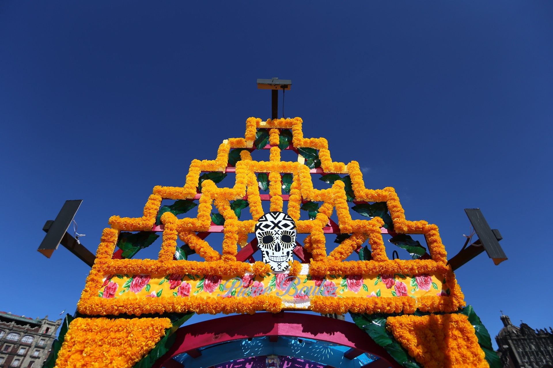 Ein Día de los Muertos-Bogen, geschmückt mit Blumen und einem Schädel, steht in Mexiko-Stadt, eingerahmt von Gebäuden unter einem klaren blauen Himmel.