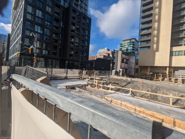 Baustelle in Toronto, Ontario mit einem großen Bauwerk im Hintergrund, umgeben von Absperrungen, Pfählen, Verkehrssignalen und Schildern unter einem bewölkten Himmel.