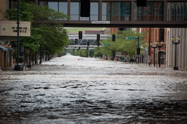Überschwemmte Stadtstraße mit Wasser, das die Straße, Pfosten, Laternen, Schilder, Verkehrszeichen, Bäume, Gebäude und eine Brücke im Hintergrund bedeckt.