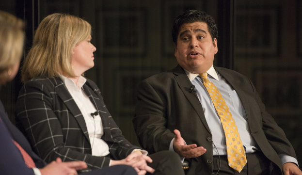 Ein Mann in einem schwarzen Blazer, weißem Hemd und gelber Krawatte sitzt neben einer Frau in einem Anzug auf einer dunklen Bühne.