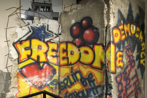 Mauer mit Graffiti, das "Freiheit und Demokratie" in fetten schwarzen Buchstaben liest, einem Geländer unten und einem Gebäude mit Fenstern im Hintergrund.