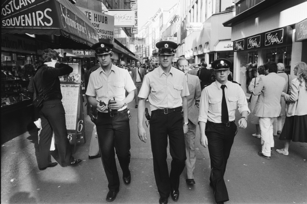 Drei Polizisten in Uniform gehen eine belebte städtische Straße mit hohen Gebäuden, Geschäften und Passanten entlang.