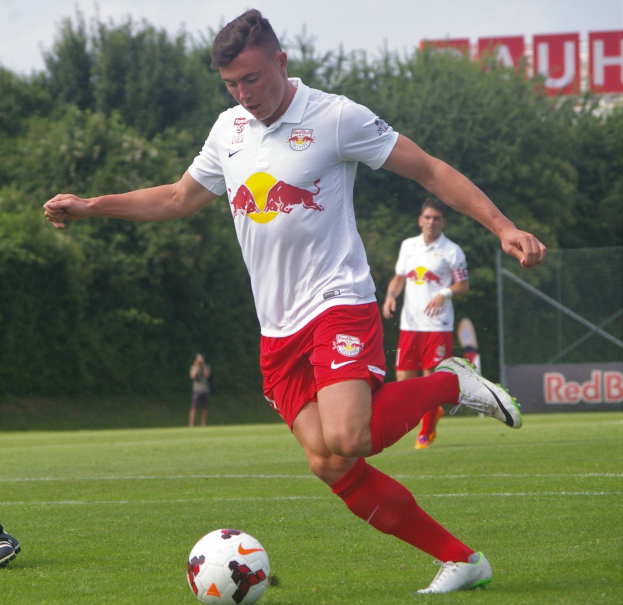 Ein Profifußballer schießt einen Ball auf einem grünen Feld mit Bäumen und einem klaren blauen Himmel im Hintergrund, mit "RB Leipzig" auf einem Schild.