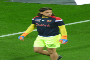 Ein Mann in einem blauen Hemd und gelben Shorts läuft auf einem Fußballfeld mit ausgestreckten Armen, entschlossen wirkend.