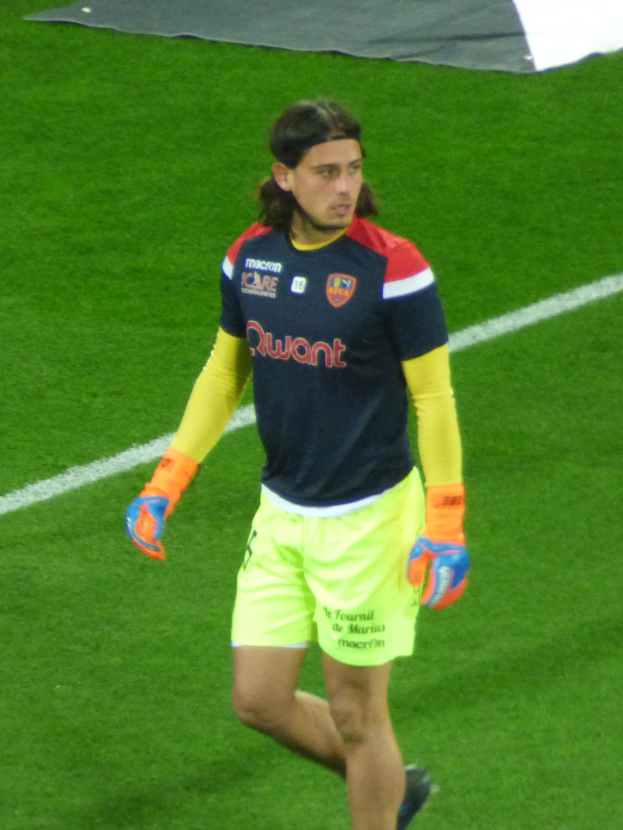 Ein Mann in einem blauen Hemd und gelben Shorts läuft auf einem Fußballfeld mit ausgestreckten Armen, entschlossen wirkend.