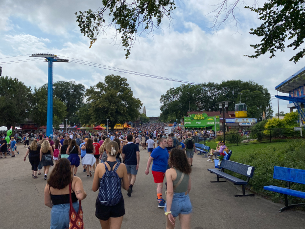 Eine Menschenmenge, die eine Straße mit Bänken, Pflanzen, Gras, Bäumen, Pfählen, Laternen, Drähten und einem Schuppen entlanggeht, wobei einige Taschen tragen, unter einem bewölkten Himmel.