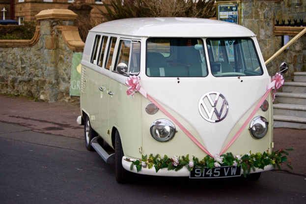 Ein wei├čer und rosafarbener VW Camper mit Blumen und Bl├Ąttern verziert, der vor einer Steinwand mit Treppe auf der rechten Seite und einem Geb├Ąude im Hintergrund geparkt ist.