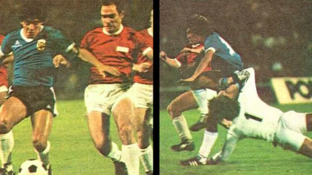 Eine Gruppe von Männern, die Fußball auf einem Feld spielen, mit einem Scoreboard im Hintergrund, ein Spieler tritt den Ball, während andere versuchen, die Kontrolle zu übernehmen.