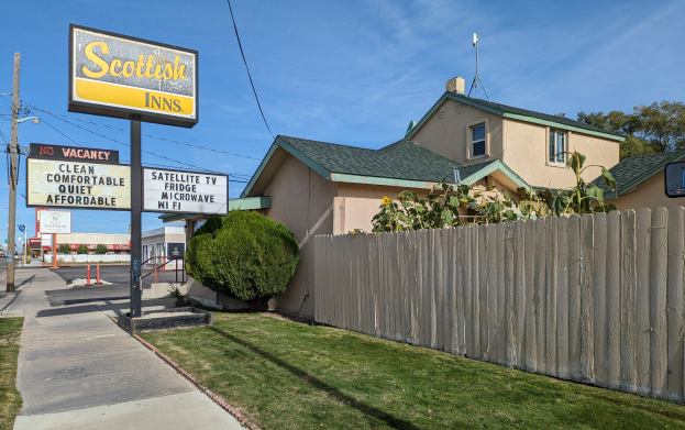 Außensicht eines schottischen Inns Motel in San Francisco, Kalifornien, das ein Haus mit Dach und Fenstern, einen Holzzaun, Pflanzen, Gras, Bäume, Schilder, Strommasten mit Drähten, eine Straßenlaterne, Fahrzeuge auf der Straße und einen bewölkten Himmel zeigt.