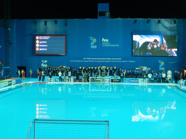 Gruppe von Menschen, die um ein Hallenbad mit Netz am Boden stehen, mit einer Wand im Hintergrund, die den Text "FINA World Championships 2015 Kazan" und Logos anzeigt.