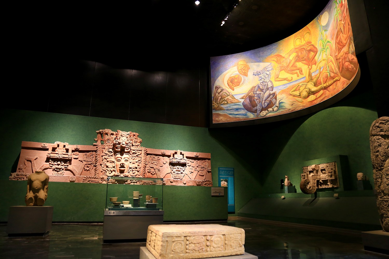 Ein Museumsraum mit verschiedenen Artefakten, darunter Skulpturen, Objekte auf Tischen, eine Texttafel, eine Wandmalerei und Deckenleuchten im Mexiko-Stadt-Museum der Anthropologie.
