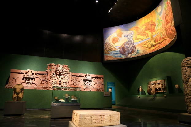 Ein Museumsraum mit verschiedenen Artefakten, darunter Skulpturen, Objekte auf Tischen, eine Texttafel, eine Wandmalerei und Deckenleuchten im Mexiko-Stadt-Museum der Anthropologie.
