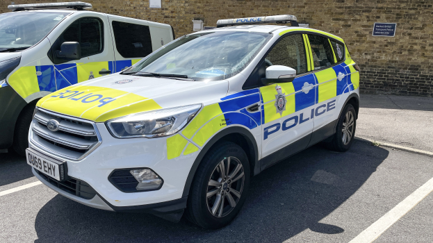 Zwei Polizeiautos nebeneinander auf einem Parkplatz geparkt, mit einer Wand und einer Schautafel im Hintergrund.