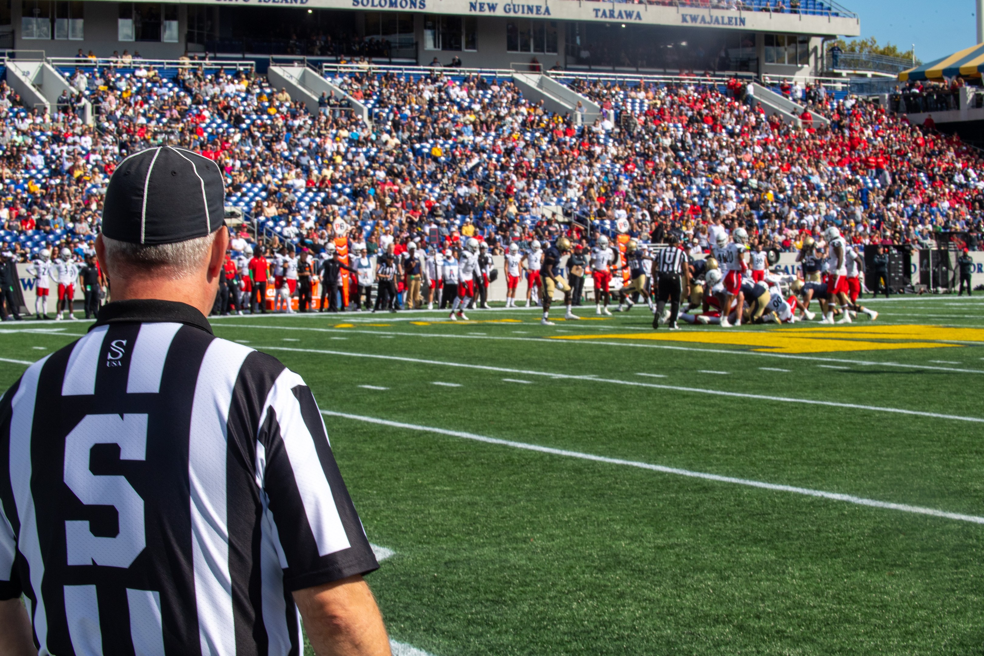 Referee standing on a football field near spectators, some wearing caps, with railings, boards, poles, trees, and a clear blue sky in the background.