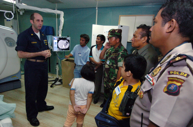 Eine Gruppe von Menschen in einem Krankenhauszimmer mit einem Mann, der ein Mikrofon hält, einem Bett mit Decke, medizinischen Geräten, einem Röntgengerät, einer Texttafel, einer Tür und Deckenleuchten.