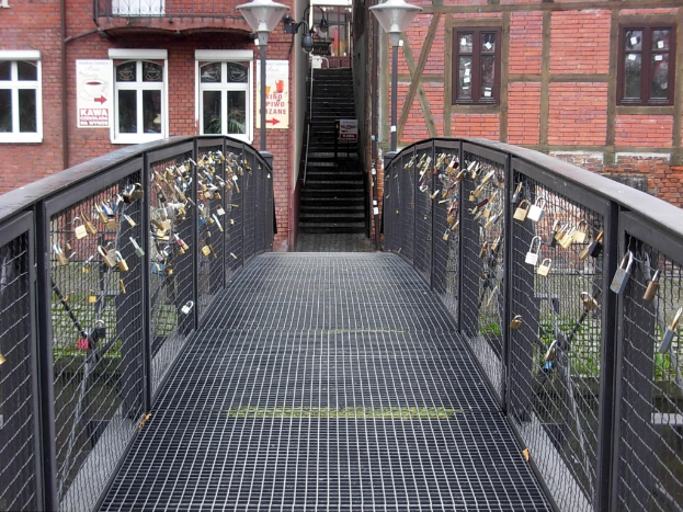 Eine Brücke, geschmückt mit Liebesloks, spannt sich vor einem Backsteingebäude, beleuchtet von Laternen, mit Stufen, die zum Gebäude führen.