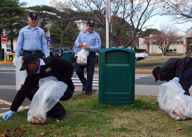 Drei Polizeibeamte in Mützen und Handschuhen, die Müll auf einer Straßenböschung mit Bäumen, Gebäuden, Schildern, Fahrzeugen und einem Mülleimer im Hintergrund, Gras und trockenen Blättern auf dem Boden und einer bewölkten Himmel sammeln.
