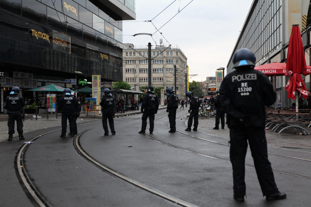 Gruppe von Polizeibeamten in schwarzen Uniformen und Helmen auf einer Straße mit Gebäuden, Bäumen und verstreuter Außenmöblierung unter einem klaren Himmel.