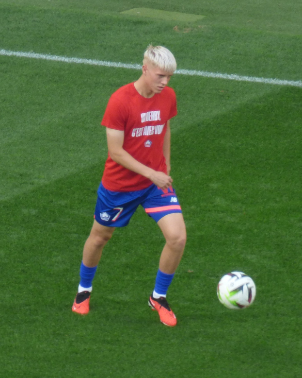 Ein junger Mann in einem roten Trikot und blauen Shorts, der auf einem grünen Rasen Fußball spielt und Fußballschuhe trägt.