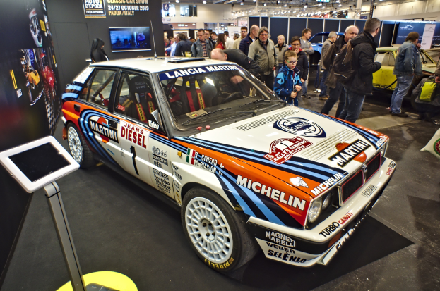 Lancia Delta HF Integrale Evoluzione Rallyefahrzeug auf einer Autauschstellung umgeben von Menschen in bunten Kleidern, mit einem Bildschirmständer links und Deckenlichtern und Tafeln im Hintergrund.
