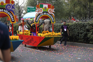Eine Gruppe von Menschen auf einem blumengeschmückten Festwagen bei einem Umzug, die verschiedene farbige Kleider und Hüte tragen, umgeben von einem Metallzaun, Schildern, einer Straßenlaterne, Bäumen und einem bewölkten Himmel.