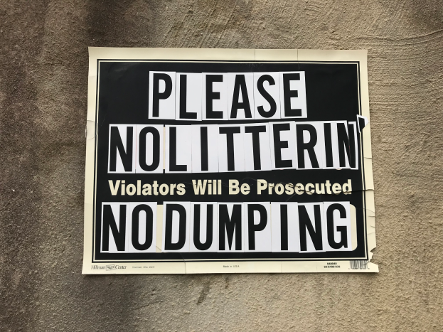 Ein schwarzes und weißes Schild an einem Gebäude trägt die Aufschrift "Bitte Nolitterin' Violators Will Be Prosecuted No Dumping".