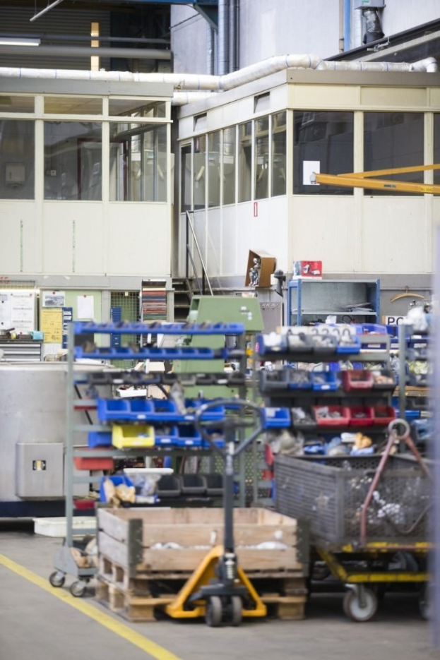 Ein großer Lagerraum voller verschiedener Geräte wie Wagen, Schränke und eine Kiste, mit einer Glaswand und Rohren im Hintergrund.