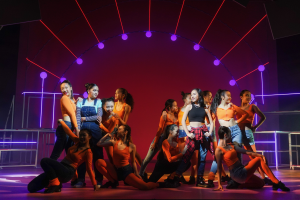 Eine Gruppe junger Tänzer in bunten Kostümen performs auf einer Bühne mit bunten Lichtern und Geländern, während das Publikum im Hintergrund jubelt.