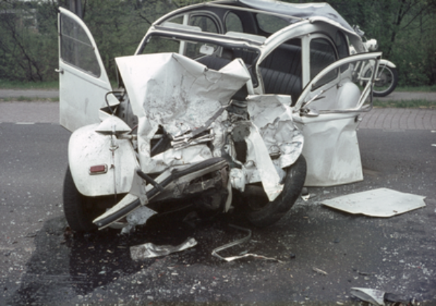 Ein stark beschädigtes Auto steht mit schwerer Frontschaden am Straßenrand, mit Bäumen, einem Gebäude und einem anderen Fahrzeug im Hintergrund.