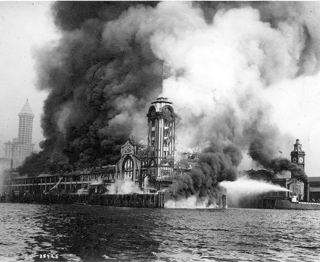 Schwarzes und weißes Foto eines großen, von Flammen eingehüllten Gebäudes mit Rauch, der herausquillt, einem Gewässer im Vordergrund und anderen Gebäuden und dem Himmel im Hintergrund.