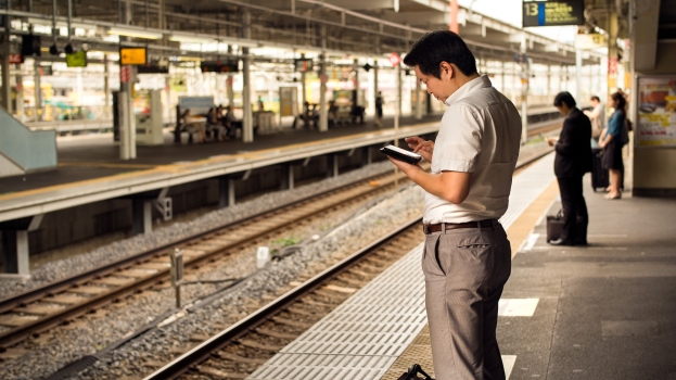 Ein Mann steht auf einem Bahnsteig und schaut auf sein Handy, umgeben von anderen Menschen, mit Bahnschienen im Hintergrund und einer Anzeigetafel auf der rechten Seite.