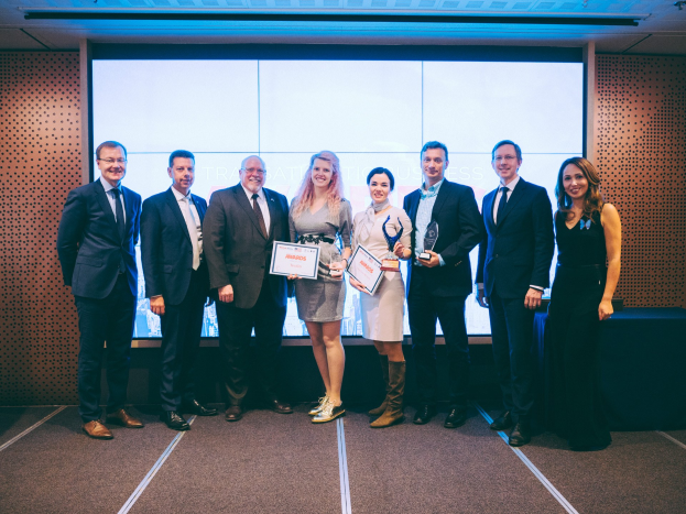 Gruppe von strahlenden Menschen mit Urkunden, die vor einem Bildschirm stehen, mit einem Tisch und einem Objekt an der Wand im Hintergrund, Gewinner der Internationalen Business Awards 2018.