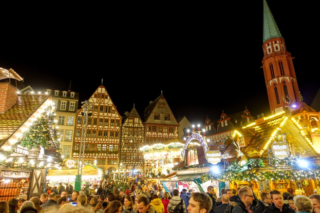 Ein lebendiger Weihnachtsmarkt in Nürnberg, Deutschland, mit Menschen um beleuchtete und dekorierte Stände, Gebäude mit Fenstern und einem Uhrturm im Hintergrund unter einem dunklen Himmel.