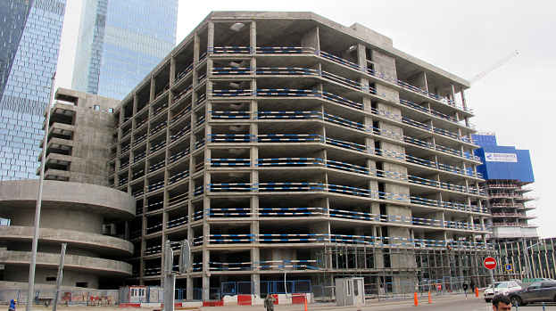 Großes Gebäude im Bau in einer Stadt umgeben von Wolkenkratzern, Fahrzeugen, Fußgängern, Verkehrsinfrastruktur und Kränen unter einem klaren blauen Himmel.