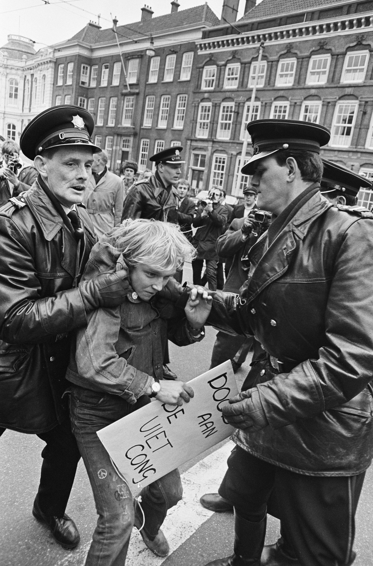 Schwarz-weißes Bild einer Frau, die von zwei Polizisten auf einer Straße eskortiert wird, mit Zuschauern und Gebäuden im Hintergrund.