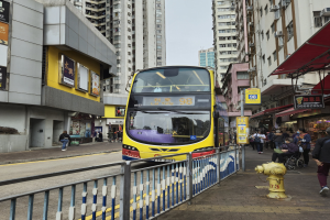 Ein gelber Doppeldeckerbus fährt eine von hohen Gebäuden gesäumte Straße entlang, mit Menschen auf dem rechten Gehweg, einem Hydranten und einem Geländer, unter einem klaren blauen Himmel.
