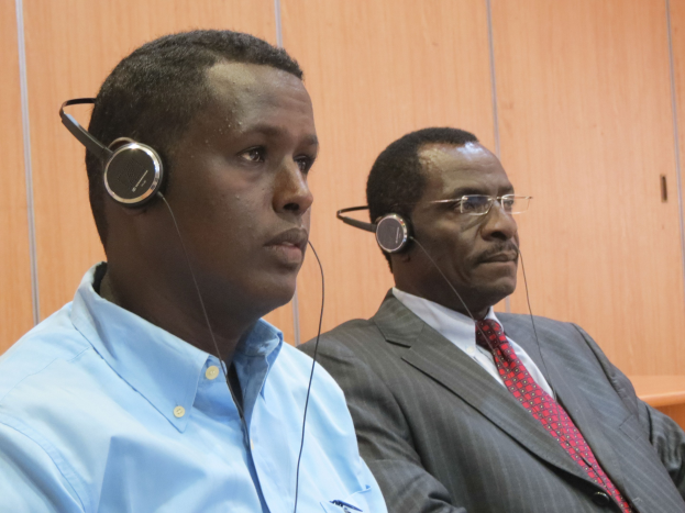 Zwei Männer in einem Gerichtssaal mit Kopfhörern, einer trägt ein blaues Hemd auf der linken Seite und einer mit Brille in der Mitte, vor einer hölzernen Wand sitzend.