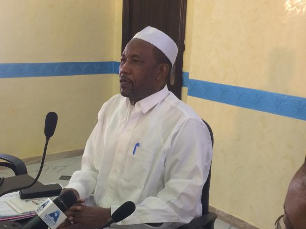 Ein Mann in einem weißen Gewand und Hut sitzt an einem Tisch mit Mikrofonen, spricht, während Papiere und Gegenstände auf dem Tisch zu sehen sind, mit einer anderen Person mit Brille zu seiner Rechten, in einem Raum mit einer Wand und einer Tür dahinter.