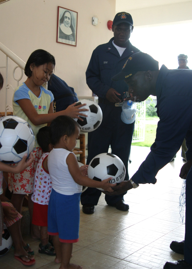 Eine Gruppe von Kindern und ein Mann mit einem Fussball, mit einer Flasche in der Hand des Mannes, der in der Nähe einer Wand mit einem Foto-Rahmen und einem Geländer steht.