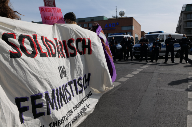 Gruppe von Menschen, die eine "Solidarität und Feminismus"-Tafel tragen, die eine Straße entlangmarschieren, mit geparkten Fahrzeugen, Gebäuden, einer Schüsselantenne und einem klaren blauen Himmel im Hintergrund.