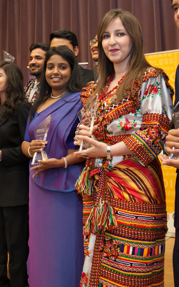 Gruppe von Menschen auf einer Bühne mit einer Frau in der Mitte, die eine Trophäe hält, vor einem Vorhang und einer Tafel mit der Aufschrift "Bravo Segen Award Gewinner."