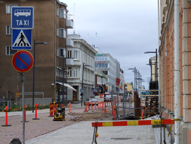 Eine Stadtstraße mit Gebäuden, Straßenlaternen, Schildern, Verkehrskegeln, Fahrzeugen, Absperrpollern, Bäumen und einer Baustelle mit Verkehrsschildern unter einem bewölkten Himmel.