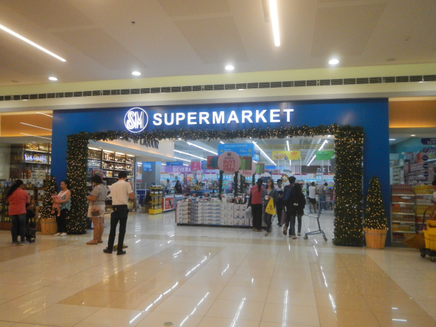 Menschen vor einem Supermarkt mit Einkaufstägern, Taschen, einem Einkaufswagen, einem Topf mit Pflanze, Regalen mit Gegenständen, Schildern und Deckenlichtern im Hintergrund.