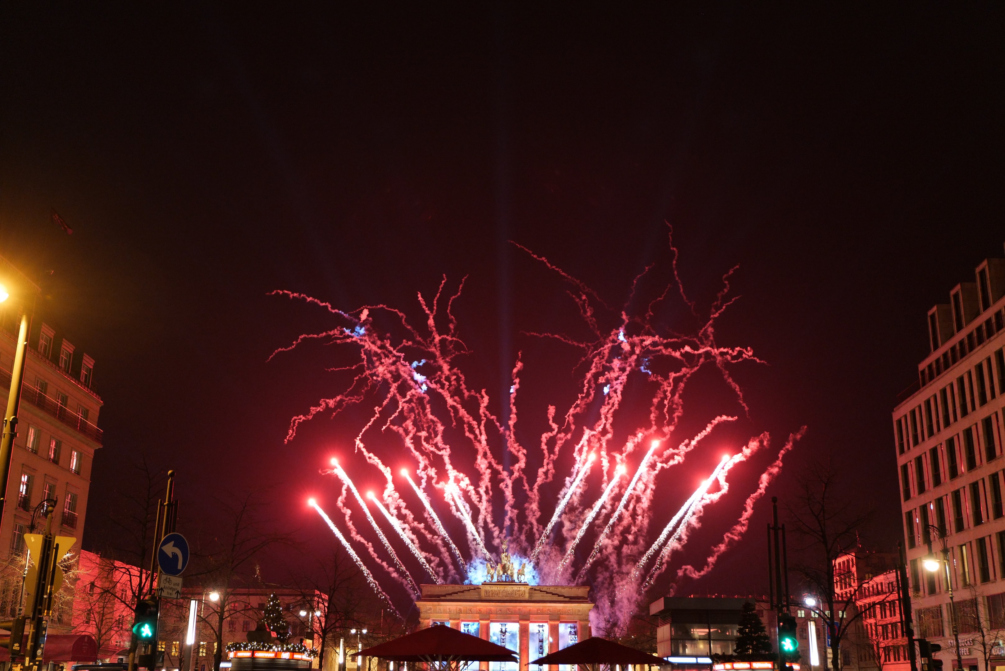 Eine belebte Stadtstraße während einer Silvesterfeier in Berlin, mit Menschenmassen, Fahrzeugen, Gebäuden und Feuerwerk, das den Nachthimmel erhellt.