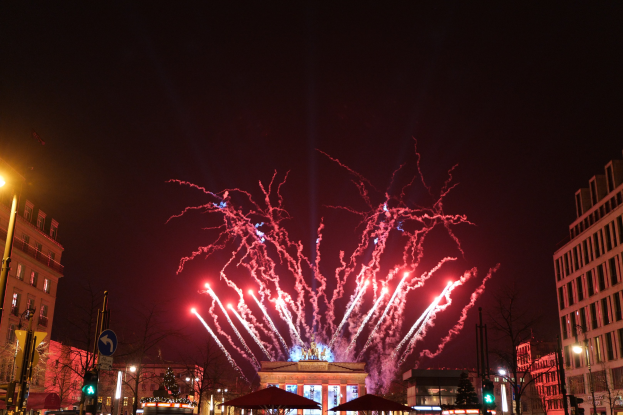 Eine belebte Stadtstraße während einer Silvesterfeier in Berlin, mit Menschenmassen, Fahrzeugen, Gebäuden und Feuerwerk, das den Nachthimmel erhellt.